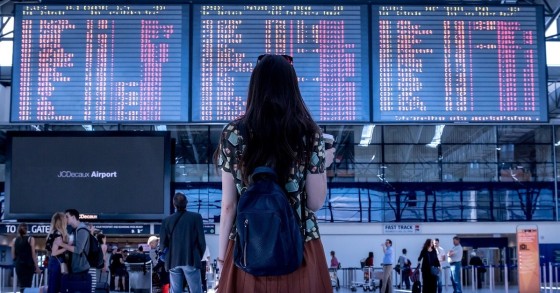 L'immagine di una ragazza con una gonna rossa ed uno zaino nero sulle spalle davanti ad un tabellone delle partenze