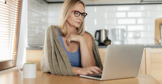 Donna utilizza un computer portatile mentre è seduta al tavolo della cucina con un'espressione pensierosa e riflessiva