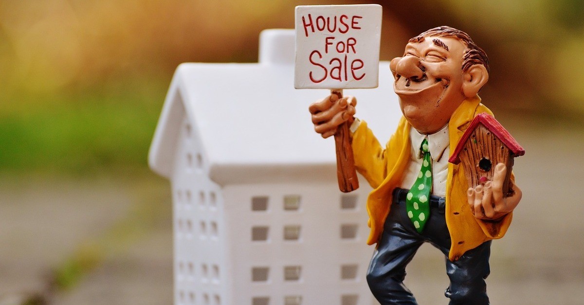 L'immagine di un piccolo uomo con giaccia arancione ed un cartello vicino ad una casa bianca