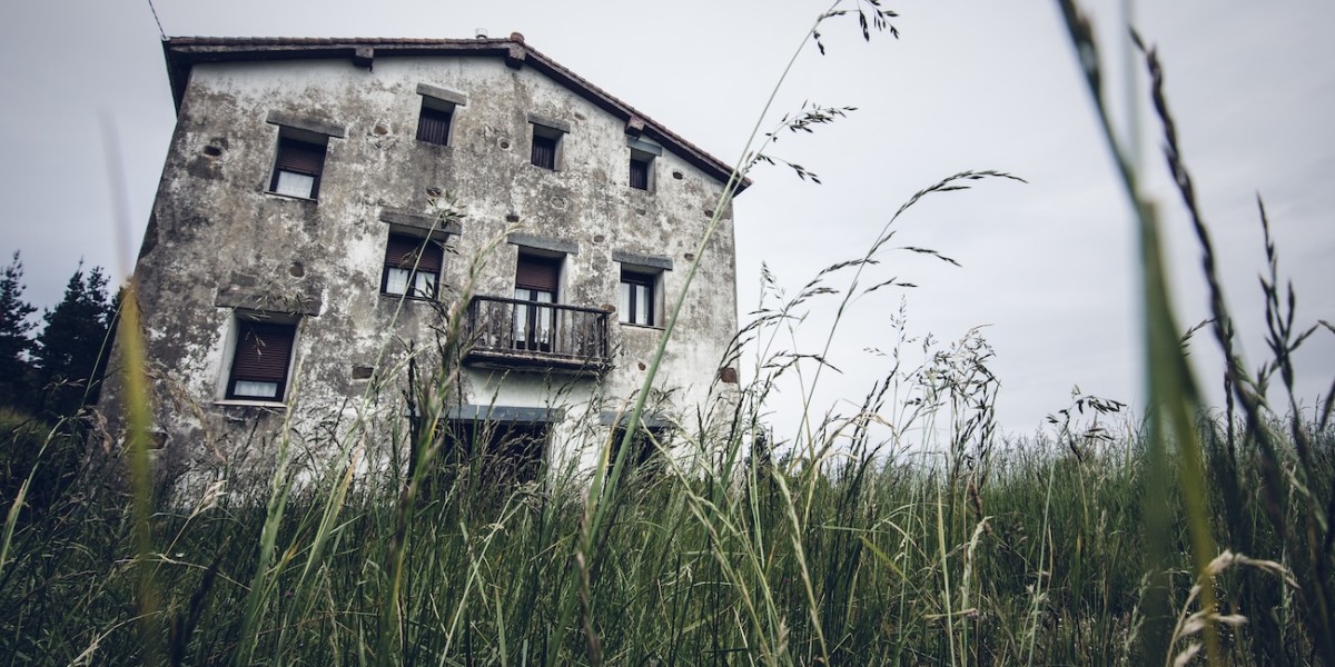 Erba molto alta, sterpaglie e una casa sullo sfondo in stato di incuria