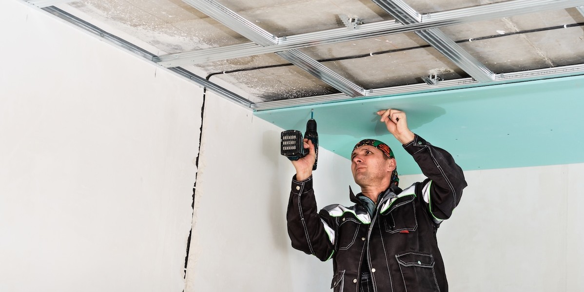 Operaio edile monta un controsoffitto in cartongesso, fissando i pannelli alla struttura metallica con un avvitatore