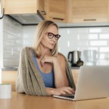 Donna utilizza un computer portatile mentre è seduta al tavolo della cucina con un'espressione pensierosa e riflessiva